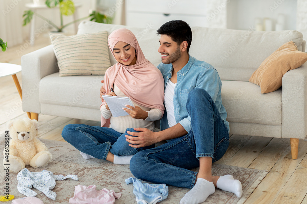 Loving pregnant muslim couple getting ready for childbirth, making checklist while sitting with baby clothes around Loving pregnant muslim couple getting ready for childbirth, making checklist while sitting with baby clothes around