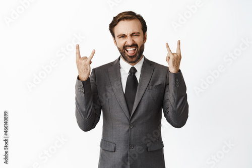 Happy corporate man shows rock on horns gesture and celebrating, smiling and rejoicing, being ecstatic, standing in suit over white background
