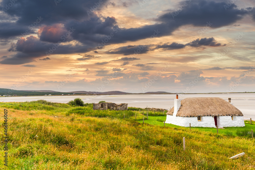 Traditionally built white cottage with thatched roof, next to the turquoise bay, with stormy cloudy dark skies above.island of North Uist, Scotland - obrazy, fototapety, plakaty 