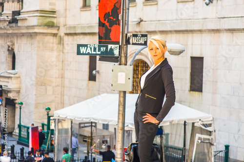 Dressing in a white underwear, black suit, carrying a bag, one hand resting on her hips, a young businesswoman is standing outside, confidently looking at you. Wall Street sign in the background.