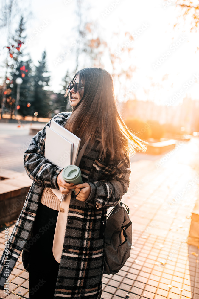 Naklejka premium Beautiful young girl student with coffee, books and a backpack spending good time at the park, music with headphones