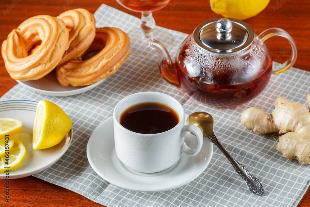 A teapot and a cup and saucer of strong aromatic tea on the table on a napkin next to ginger and lemon jam and donuts.