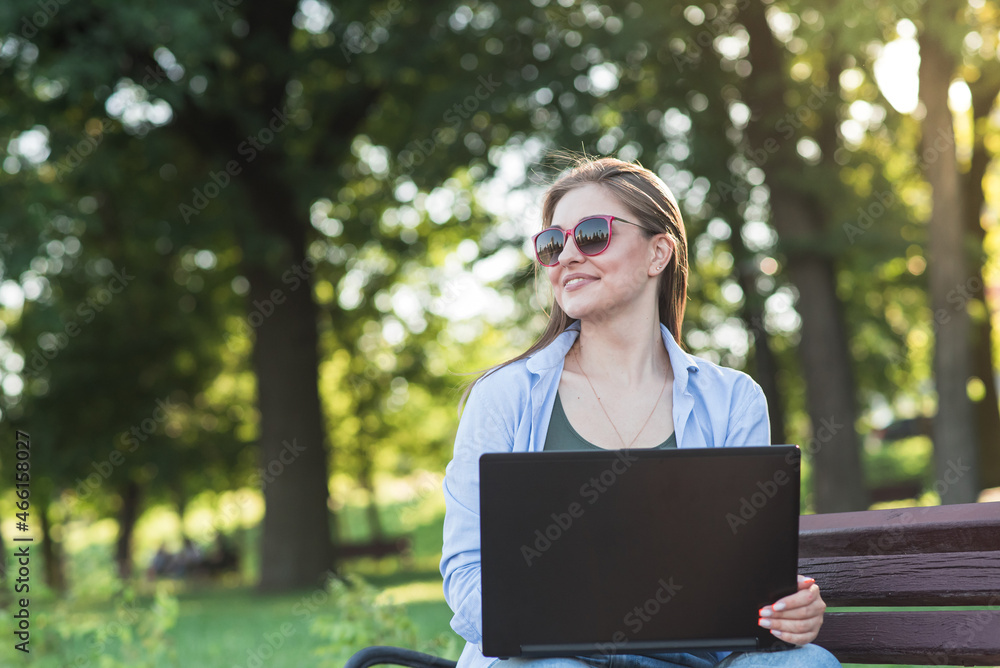 A young attractive Caucasian girl sitting on a bench in the park and using a laptop. Freelancer, student. Network concept A young attractive Caucasian girl sitting on a bench in the park and using a laptop. Freelancer, student. Network concept