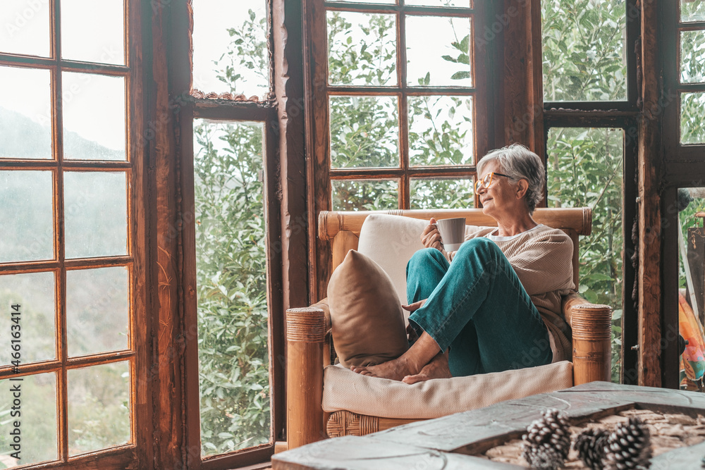 Senior woman sitting on sofa and holding coffee mug while looking away at home. Elderly woman relaxing on couch in living room. Thoughtful woman having coffee on sofa at home.