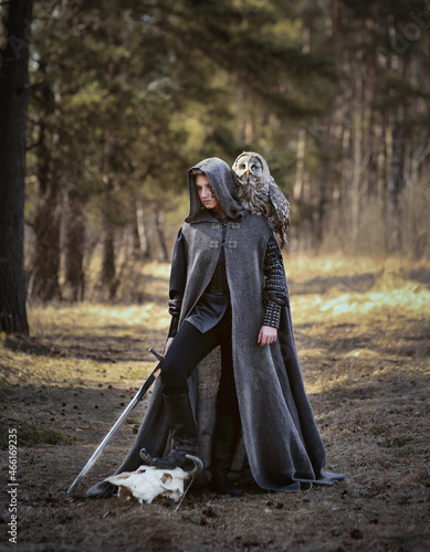 Male warrior in grey cloak and long sword in hand with owl on his shoulder standing in the forest. Fantasy portrait of a youn man in warrior costume