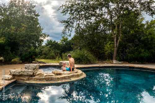 A mature man and woman enjoy a dip in a spa connected, above, a swimming pool and a waterfall, Hill Country, Texas