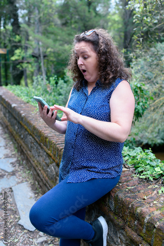 Caucasian woman, hair pulled back with a pair of sunglasses and a surprised face looking at the smartphone in a forest while resting leaning against it.