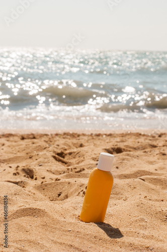 A yellow jar with a white cap with sunscreen stands in the sand on a yellow sandy beach against the background of a blue sea in a vertical format