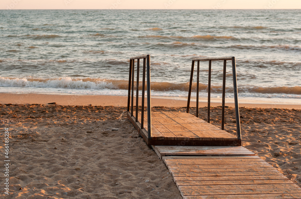 A wooden bridge with a metal railing and a wooden deck on a sandy beach leading to the sea coast at sunset A wooden bridge with a metal railing and a wooden deck on a sandy beach leading to the sea coast at sunset