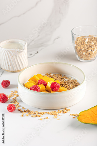 Bowl of granola with yogurt  milk and fresh raspberries  blueberries  mango for a healthy breakfast on a white marble table