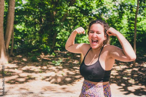 Woman in sportswear making strength gesture and smiling. Young woman in gym outfit outdoors making strength and victory gesture smiling. Photo with space for text. 