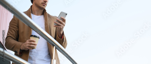 Low angle view of businessman in coat using smartphone and holding coffee to go near railing with blue sky at background, banner