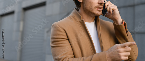 Cropped view of businessman in coat talking on smartphone and pointing with finger on urban street, banner 