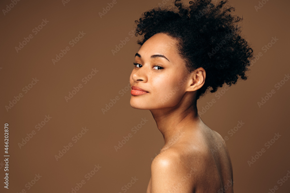 Fashion portrait of the gorgeous multiracial woman with nude makeup looking at the camera from her shoulder isolated on beige background