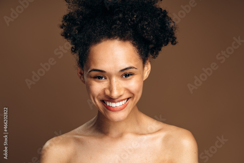 Portrait view of the multiracial woman smiling toothy and posing to the camera while standing in front of the brown wall. Stock photo