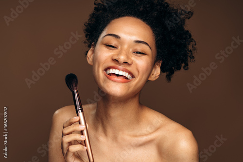 Waist up photo of a young woman looking relaxed and smiling while holding brush for makeup. Beauty procedures concept