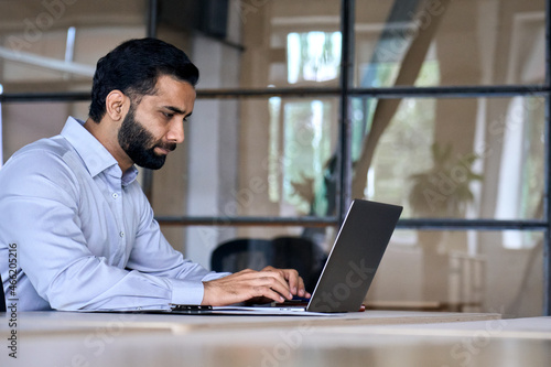 Side view of serious adult concentrated focused Indian Hispanic boss ceo businessman using typing on computer pc laptop working in contemporary office  accounting analysing report financial data.
