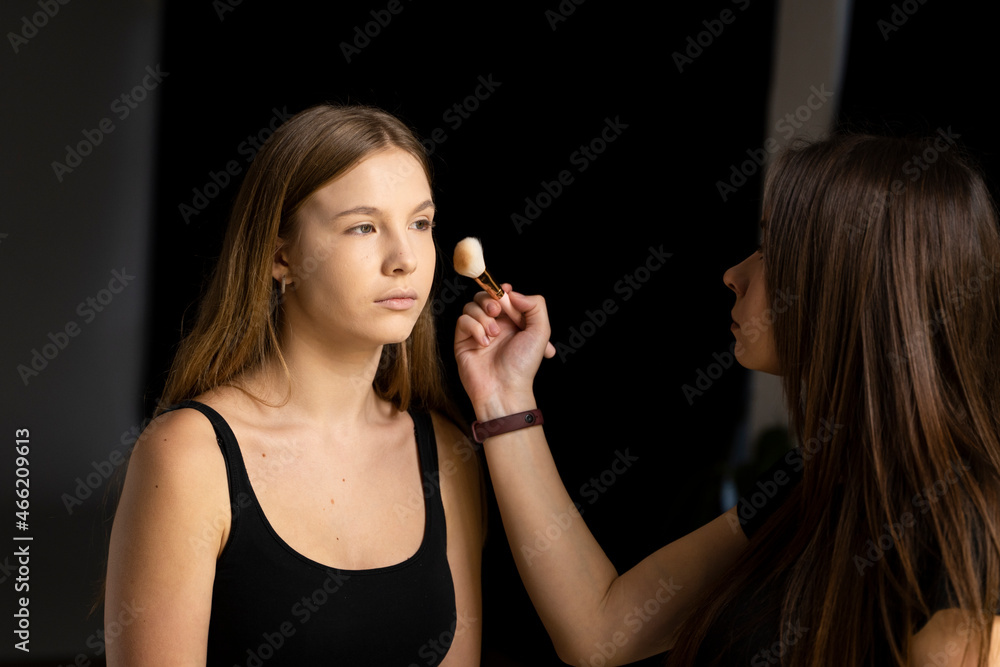 Professional make-up artist working with brush on model face. Process of making makeup. Portrait of young blonde woman in beauty salon interior. Applying tone to skin.