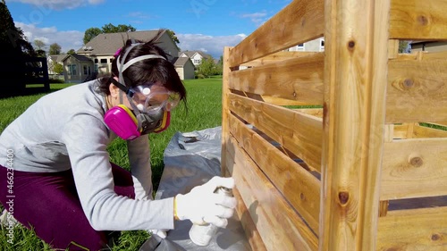 A young caucasian woman wearing face mask safety goggles and gloves is spray staining  a wooden fence outdoors. Concept video for diy project and health and safety measures she wears gas mask.