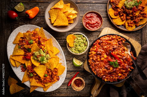 Mexican food concept: tortilla chips, guacamole, salsa, chilli con carne and fresh ingredients over wooden background, top view