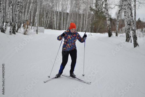 A young girl in a motley dark jacket and an orange hat goes cross-country skiing. Snowy winter in Russia. Outdoor sports and healthy lifestyle. Endurance and prevention of colds, immunity.