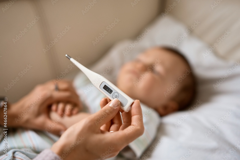 Worried young mother sitting on sofa beside her sick son with high fever. Mom measures temperature using thermometer of sick child lying under blanket at home. Worried young mother sitting on sofa beside her sick son with high fever. Mom measures temperature using thermometer of sick child lying under blanket at home.