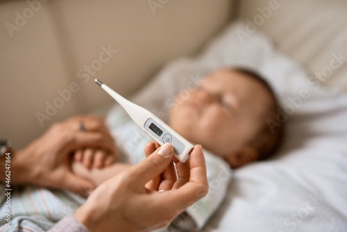 Worried young mother sitting on sofa beside her sick son with high fever. Mom measures temperature using thermometer of sick child lying under blanket at home.