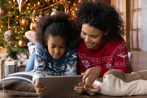 Happy biracial young mother and small daughter enjoy Christmas winter holidays using tablet gadget together. Smiling African American mom and little girl child relax at home playing on pad device. © fizkes