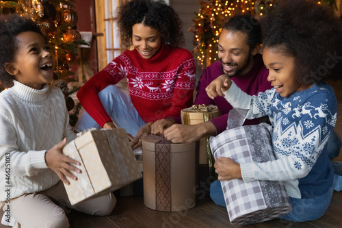 Overjoyed young African American family with two small kids have fun unpack wrapped Christmas gifts together. Excited biracial parents with little children open presents on New Year holidays.