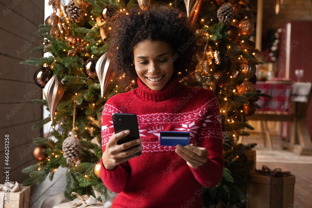 © fizkes - Happy African American woman do shopping use smartphone and credit card on New Year winter season sales. Smiling biracial female order online on cellphone, buy on Christmas discounts on web.