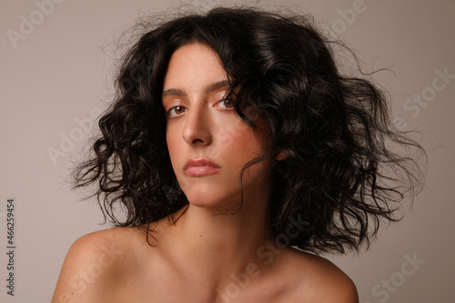 Woman with nude make-up, curly hair and naked shoulders posing over white wall.