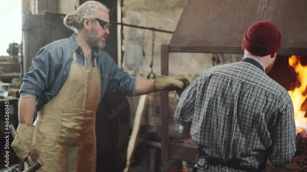 Blacksmith taking off hot iron rod from fire and giving it to senior colleague for hammering on anvil while working together in forge Blacksmith taking off hot iron rod from fire and giving it to senior colleague for hammering on anvil while working together in forge