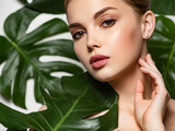 Portrait of a beautiful young girl with clean and healthy skin. Clean face of a white young girl close-up. Beautiful woman touches her face with her hand. Model with green leaves near the body. Beauty