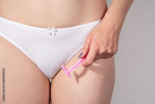 Closeup of a girl in white underwear holding a pink depilation razor in her hands on a gray background