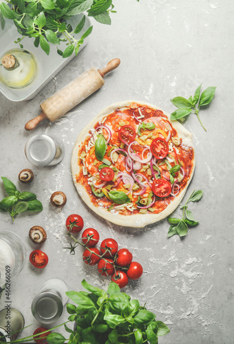 Homemade pizza with onion, salami, tomatoes and basil on grey kitchen table with wooden rolling pin, mushrooms and kitchen utensils. Preparing italian food at home with fresh ingredients. Top view.