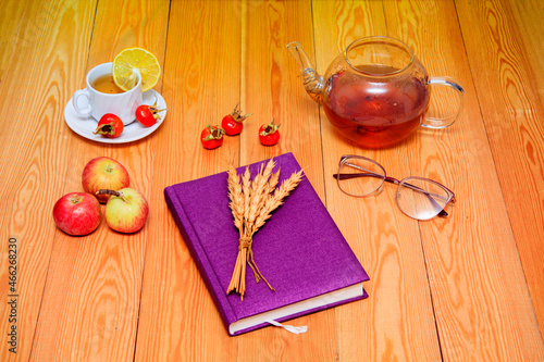 A white cup of tea with lemon.Blue notepad for notes. Glasses for vision. Wheat spikelets are tied in a bouquet. Red Apples. Glass teapot. Still-life. on a wooden background. close-up. 
