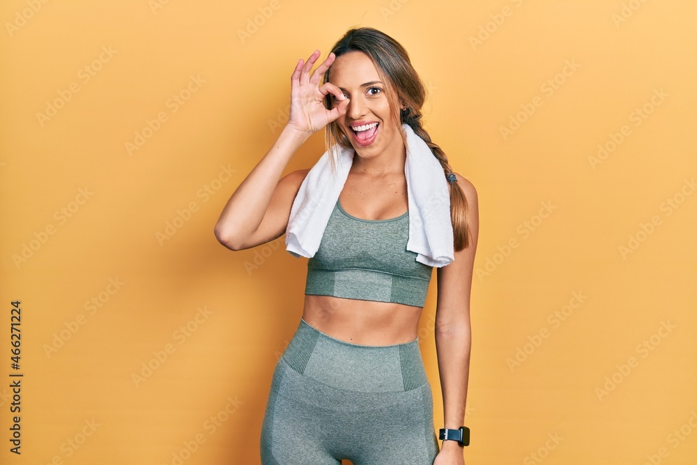 Beautiful hispanic woman wearing sportswear and towel doing ok gesture with hand smiling, eye looking through fingers with happy face. Beautiful hispanic woman wearing sportswear and towel doing ok gesture with hand smiling, eye looking through fingers with happy face.