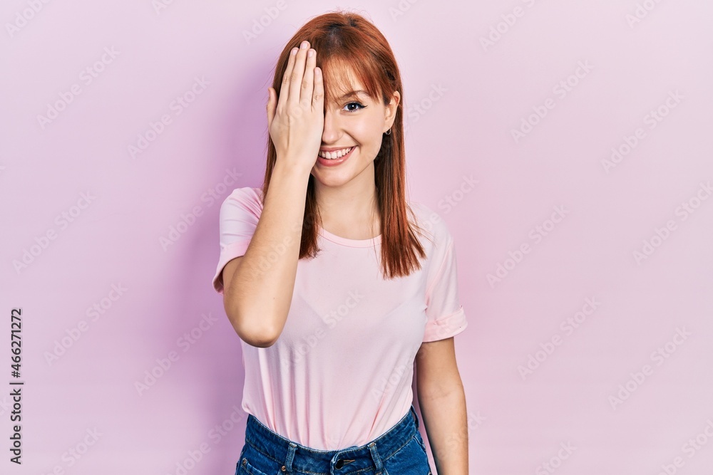 Redhead young woman wearing casual pink t shirt covering one eye with hand, confident smile on face and surprise emotion. Redhead young woman wearing casual pink t shirt covering one eye with hand, confident smile on face and surprise emotion.