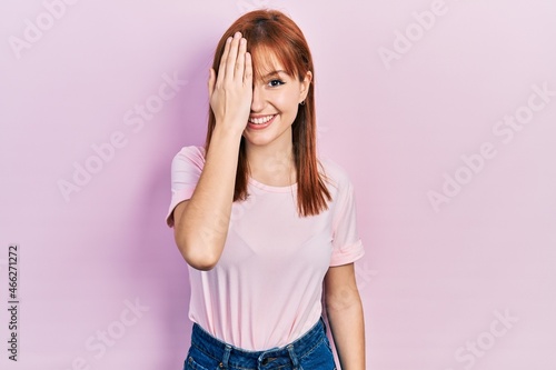 Redhead young woman wearing casual pink t shirt covering one eye with hand, confident smile on face and surprise emotion.