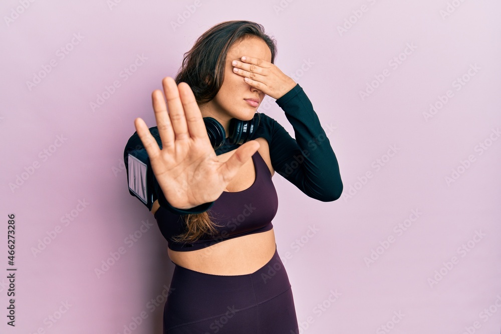 Young hispanic girl wearing sportswear and headphones covering eyes with hands and doing stop gesture with sad and fear expression. embarrassed and negative concept.