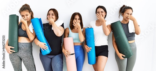 Group of women holding yoga mat standing over isolated background smelling something stinky and disgusting, intolerable smell, holding breath with fingers on nose. bad smell