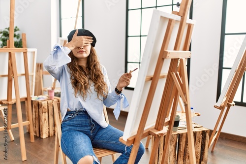Young hispanic artist woman painting on canvas at art studio covering eyes with hand, looking serious and sad. sightless, hiding and rejection concept