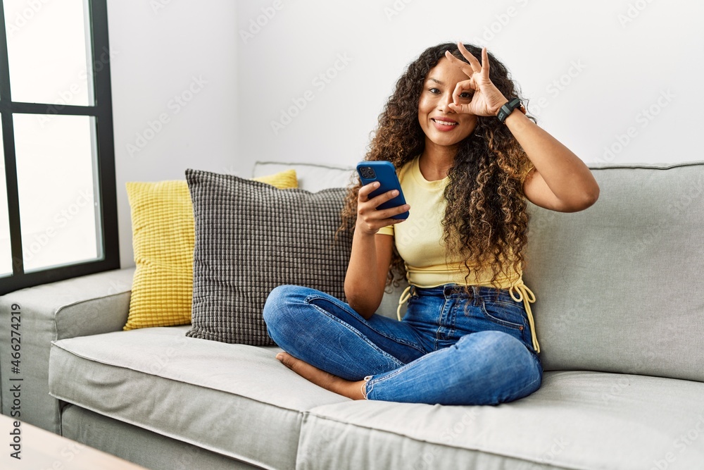 Beautiful hispanic woman sitting on the sofa at home using smartphone doing ok gesture with hand smiling, eye looking through fingers with happy face. Beautiful hispanic woman sitting on the sofa at home using smartphone doing ok gesture with hand smiling, eye looking through fingers with happy face.