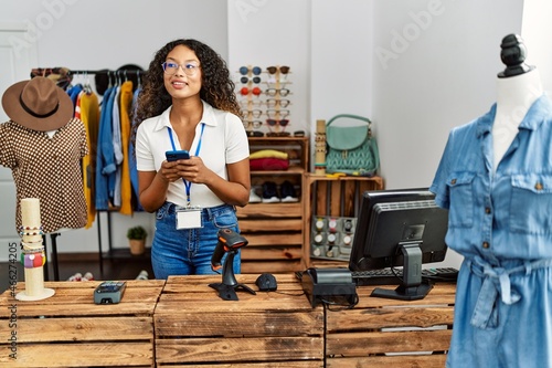 Young latin shopkeeper woman using smartphone working at clothing store.