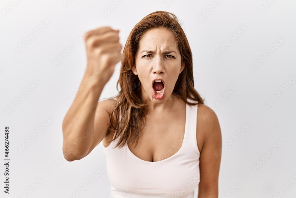 Middle age hispanic woman standing over isolated background angry and mad raising fist frustrated and furious while shouting with anger. rage and aggressive concept. Middle age hispanic woman standing over isolated background angry and mad raising fist frustrated and furious while shouting with anger. rage and aggressive concept.