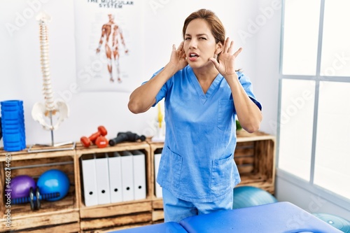 Middle age hispanic physiotherapist woman working at pain recovery clinic trying to hear both hands on ear gesture  curious for gossip. hearing problem  deaf