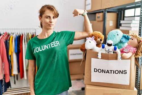 Beautiful caucasian woman wearing volunteer t shirt at donations stand strong person showing arm muscle, confident and proud of power