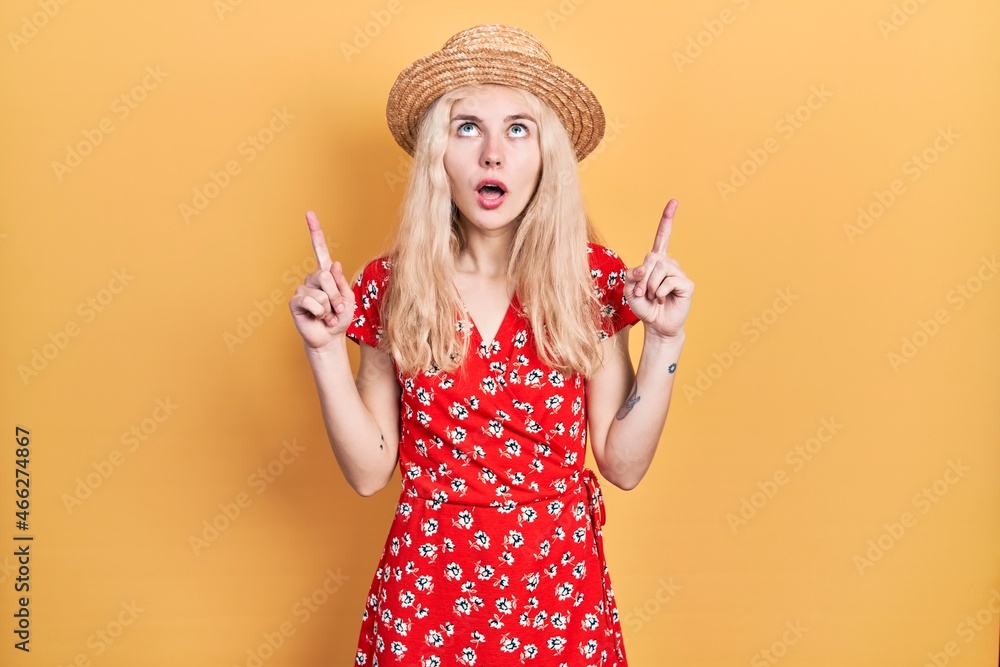 Beautiful caucasian woman with blond hair wearing summer hat amazed and surprised looking up and pointing with fingers and raised arms. Beautiful caucasian woman with blond hair wearing summer hat amazed and surprised looking up and pointing with fingers and raised arms.