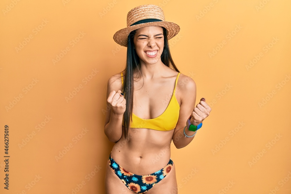 Young hispanic woman wearing bikini and summer hat excited for success with arms raised and eyes closed celebrating victory smiling. winner concept. Young hispanic woman wearing bikini and summer hat excited for success with arms raised and eyes closed celebrating victory smiling. winner concept.