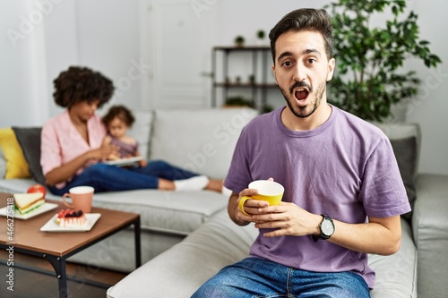 Hispanic father of interracial family drinking a cup coffee in shock face  looking skeptical and sarcastic  surprised with open mouth